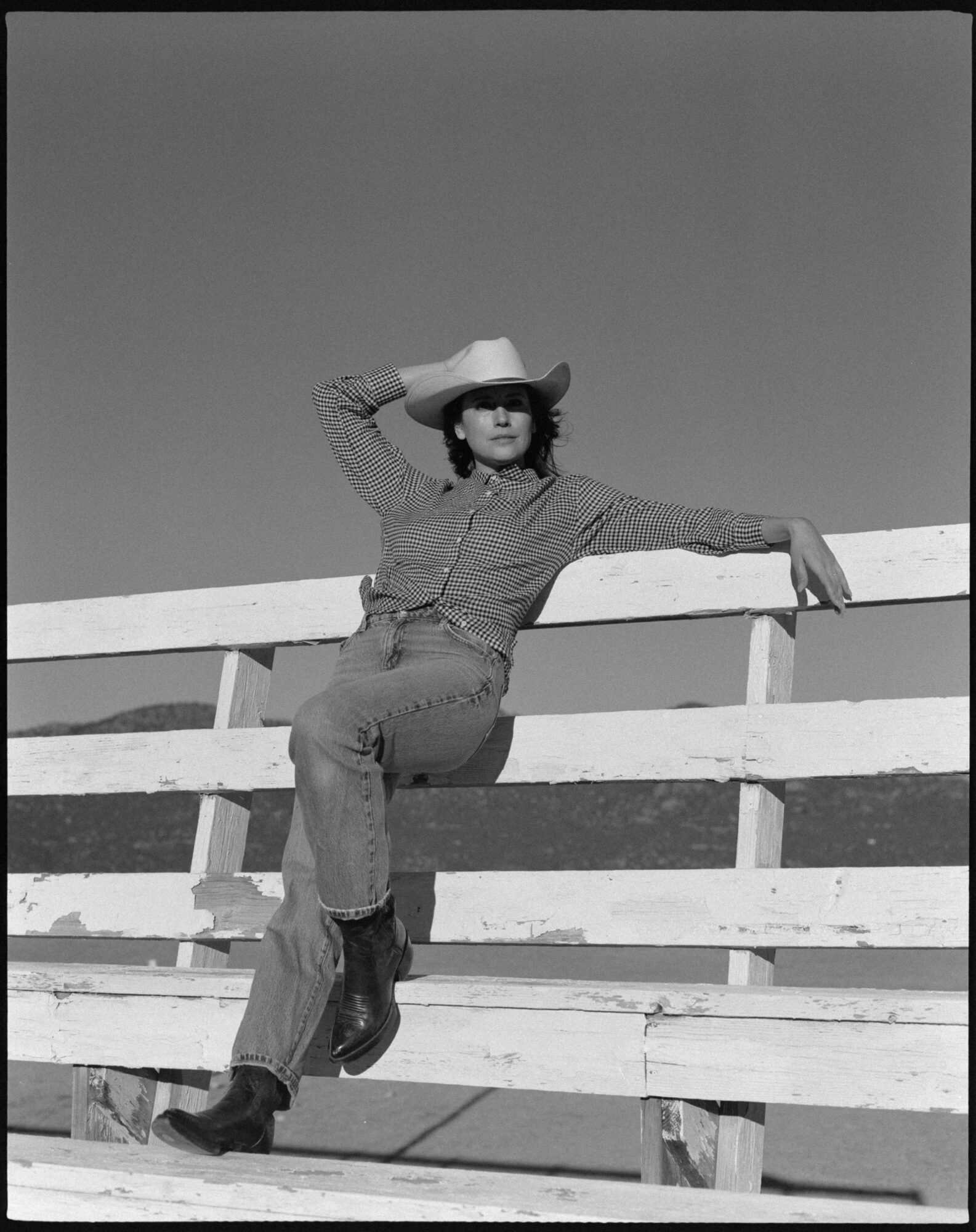 Woman in cowboy hat leaning on a wooden fence outdoors, with clear sky background.