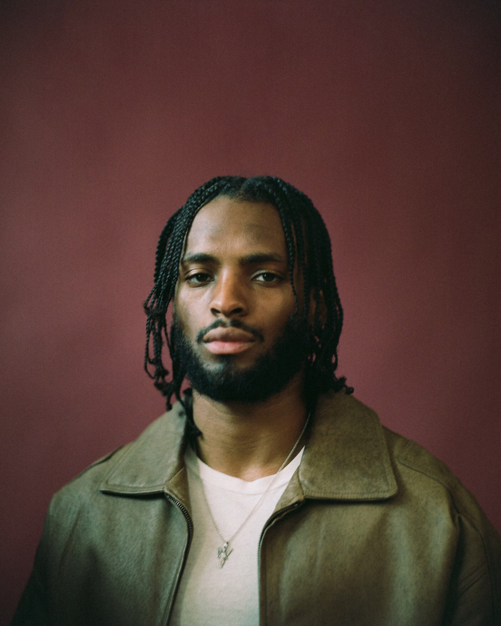 Portrait of a young man with braided hair, wearing a brown jacket and white shirt, against a dark red background.