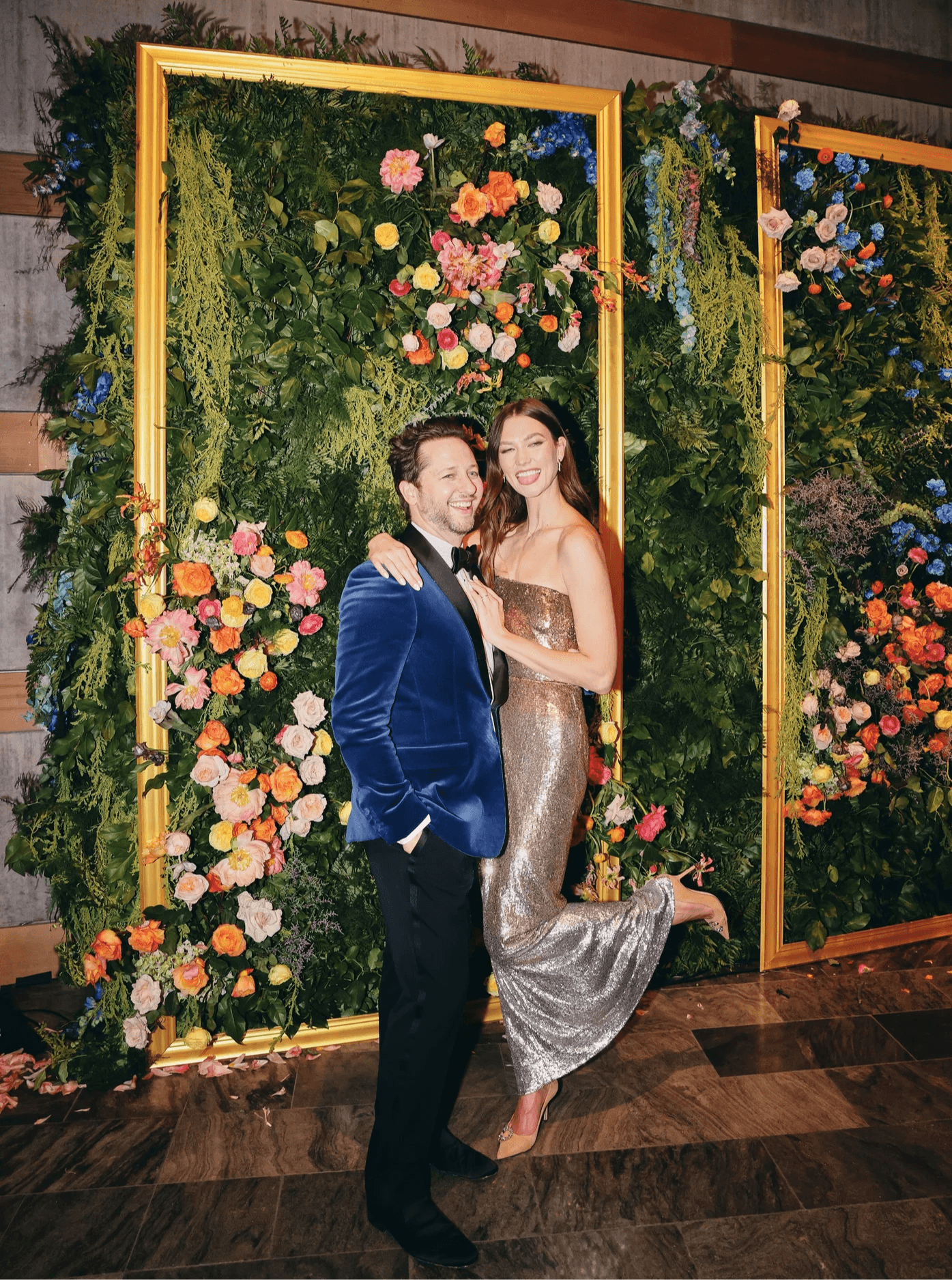 A man and woman pose in front of a floral backdrop at an event, smiling and embracing each other.