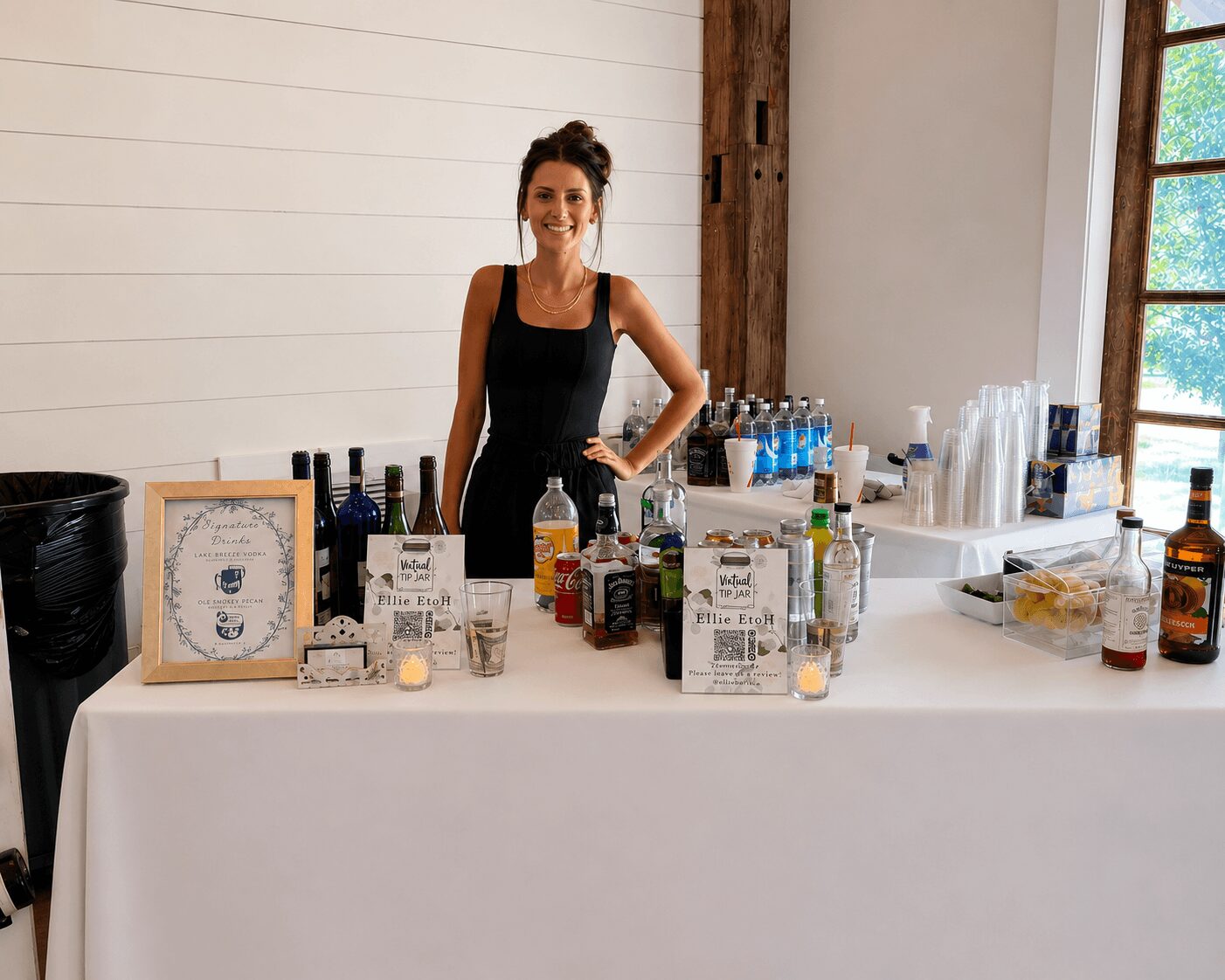 Woman standing behind a bar table with bottles and glasses, smiling, in a bright room with a window.