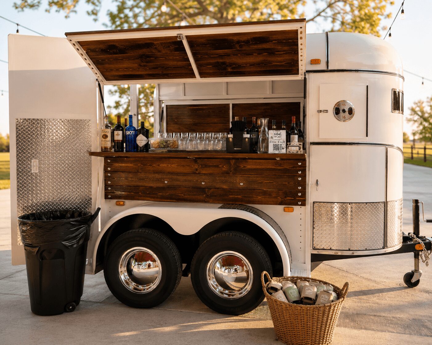 Mobile food trailer with open wooden serving window, bottles, glasses, and a basket of supplies outside, in a park setting.