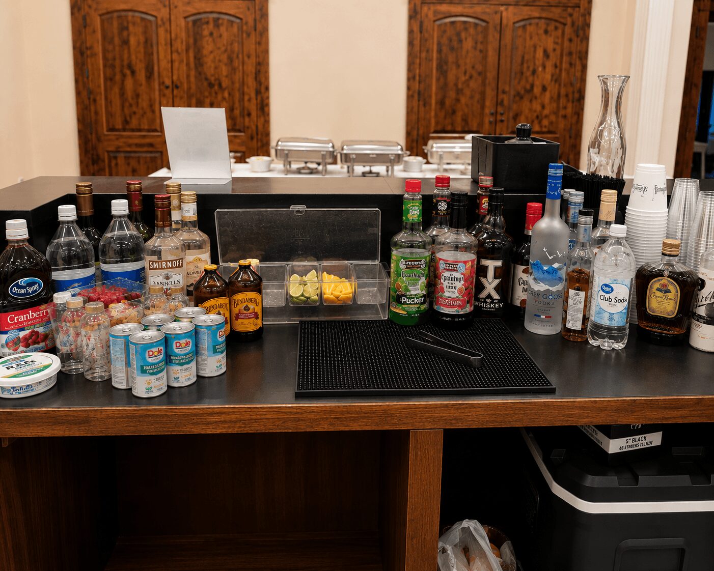 Bar with bottles, cans, and mixers on a wooden table, with two wooden doors in the background.