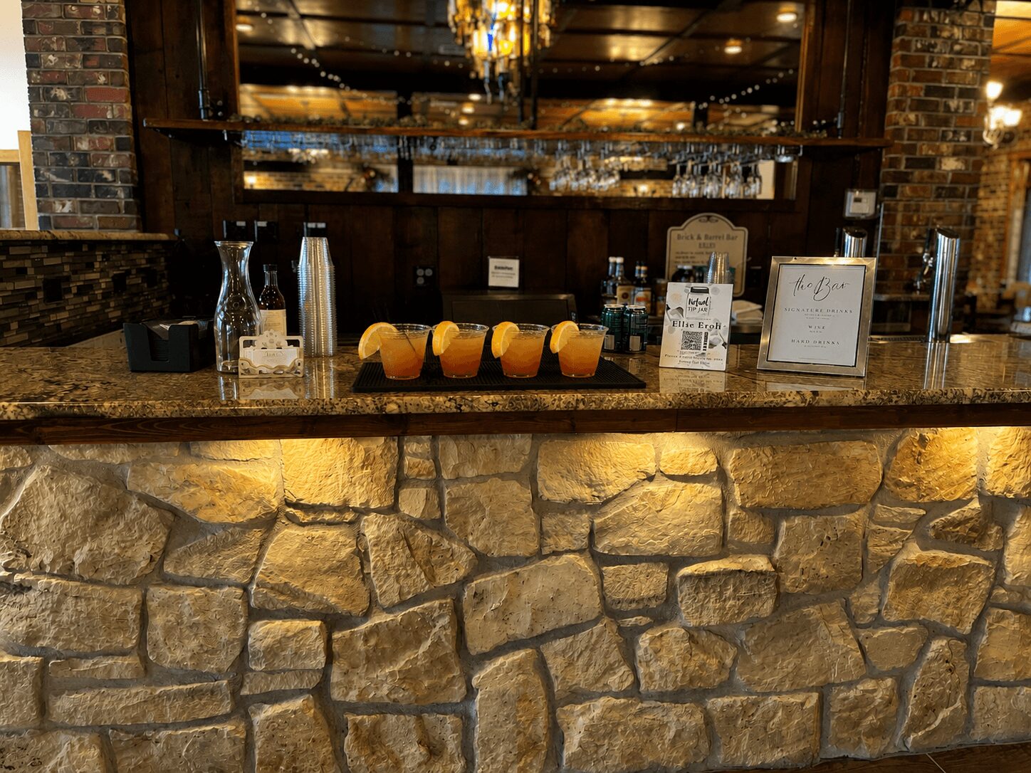 Stone bar counter with drinks and orange slices, brick walls, and a chandelier in the background.