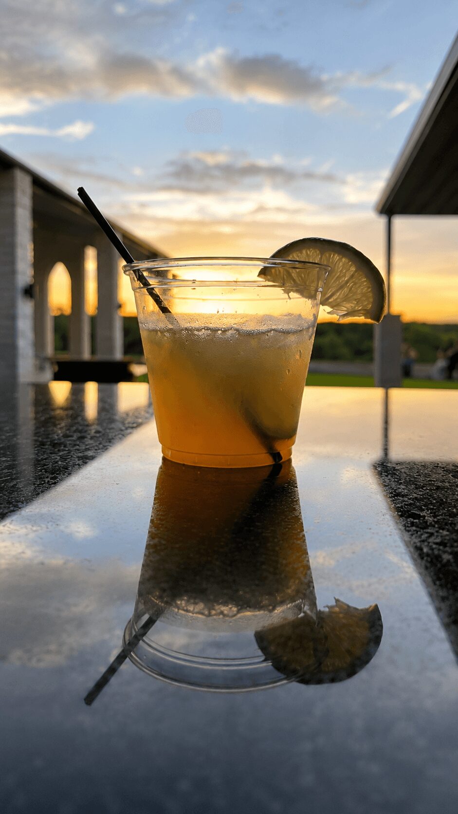 A cocktail with a lime wedge and straw on a reflective surface during sunset, with a cloudy sky and outdoor setting.