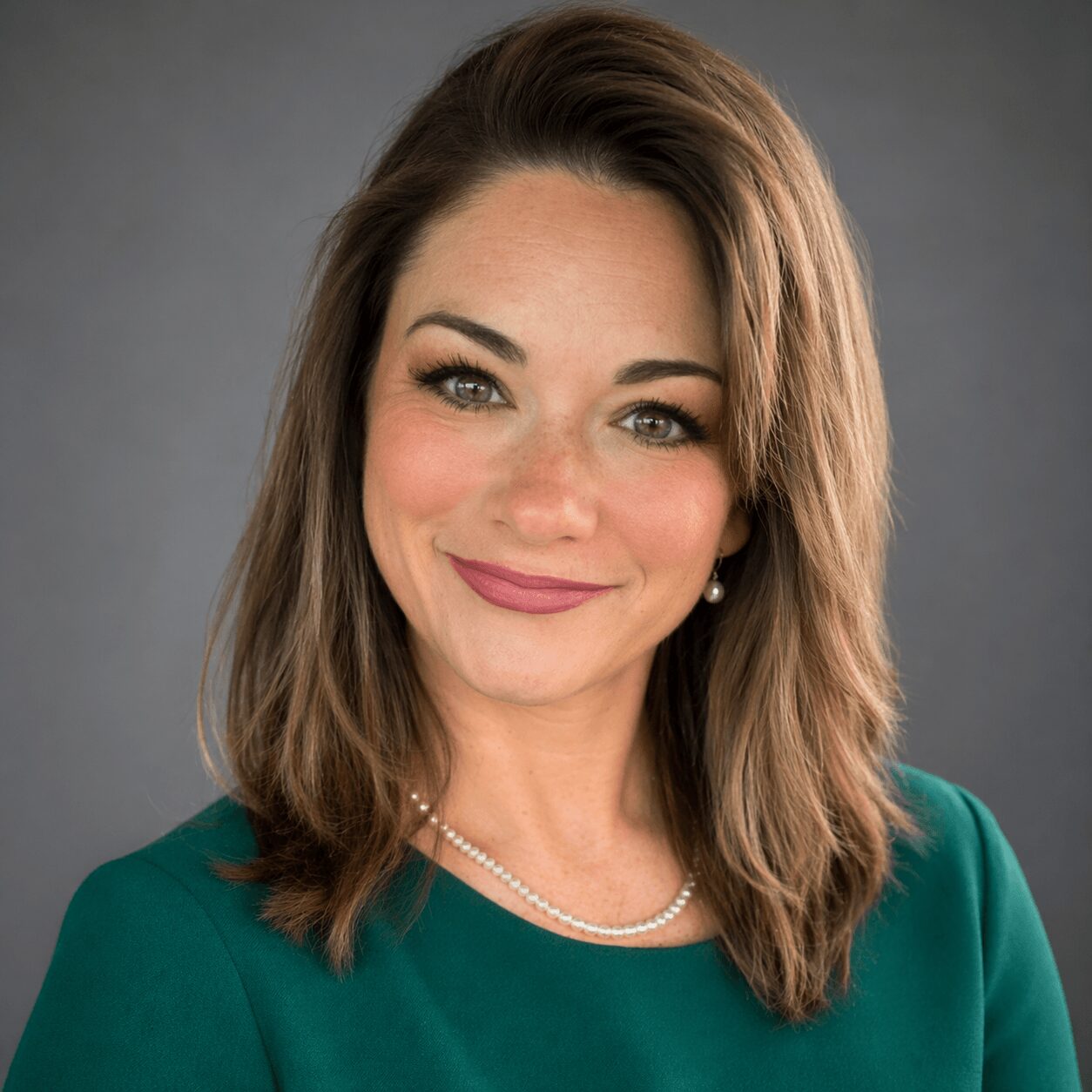 Portrait of a woman with shoulder-length brown hair, wearing a green top and pearl necklace, smiling against a gray background.