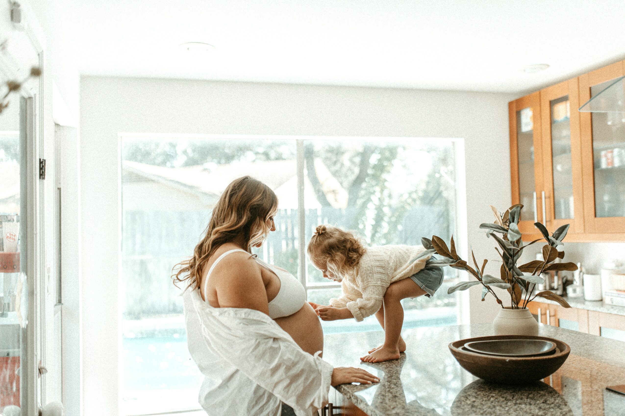 Pregnant woman with long hair in a white shirt holding her belly, a child reaching towards her, in a bright kitchen.