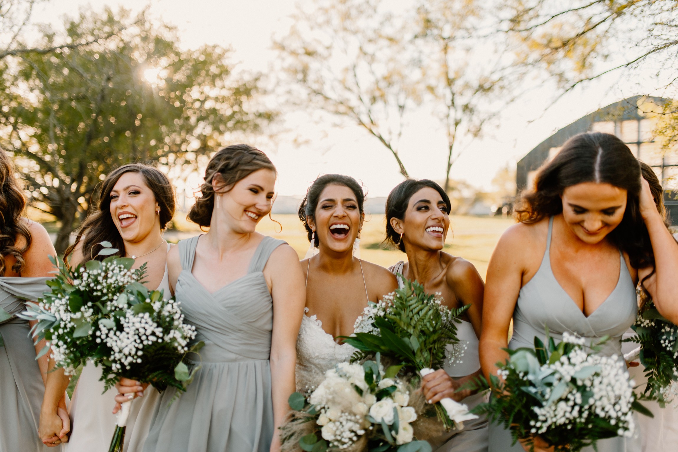 Group of women in dresses holding bouquets, smiling outdoors with trees and sunlight in background.