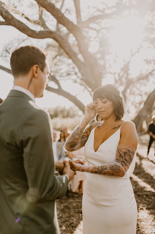 A woman in a white dress and tattoos on her arms holds hands with a man in a suit outdoors, with trees in the background.