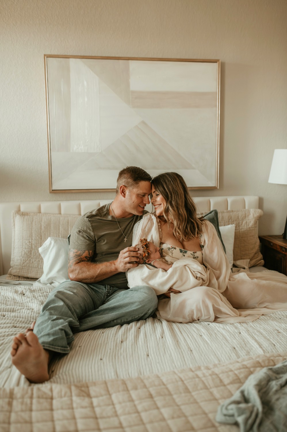 A couple sitting on a bed, holding hands and leaning close, in a softly lit room with a large framed artwork behind them.