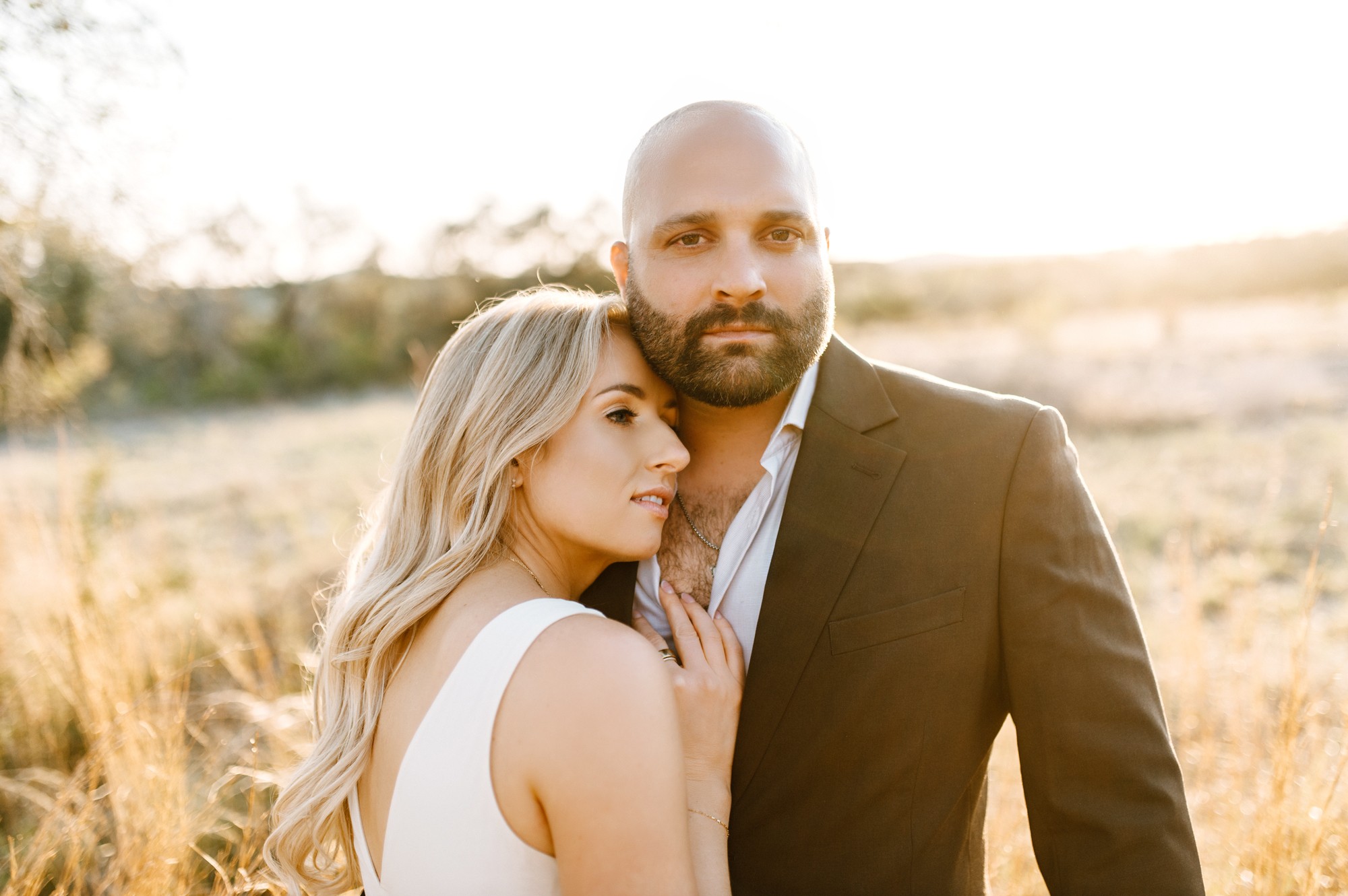 A man and woman stand close outdoors in warm sunlight, with a blurred natural background.