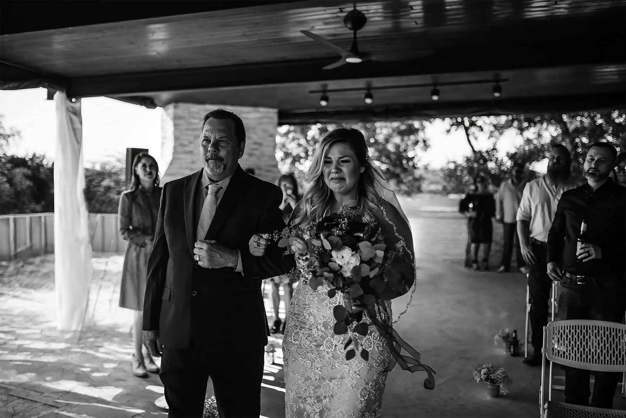 Two people walking together, one holding a bouquet, with others in the background under a covered outdoor area.