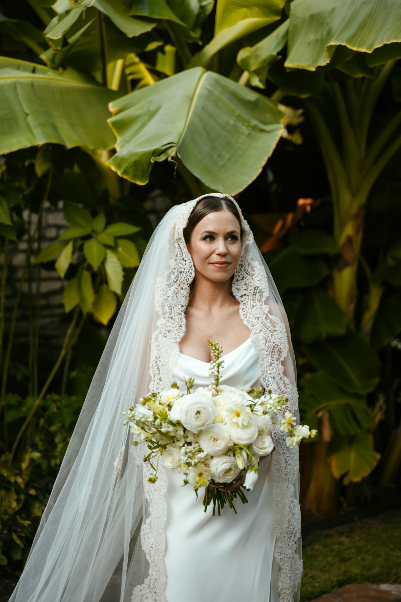 Bride in a white wedding dress holding a large bouquet of white flowers, standing among lush green banana leaves.