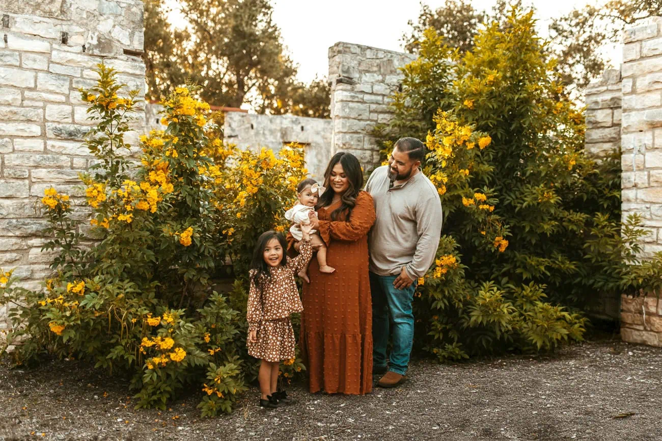 Family of four standing outdoors with yellow flowering bushes and stone walls in background.