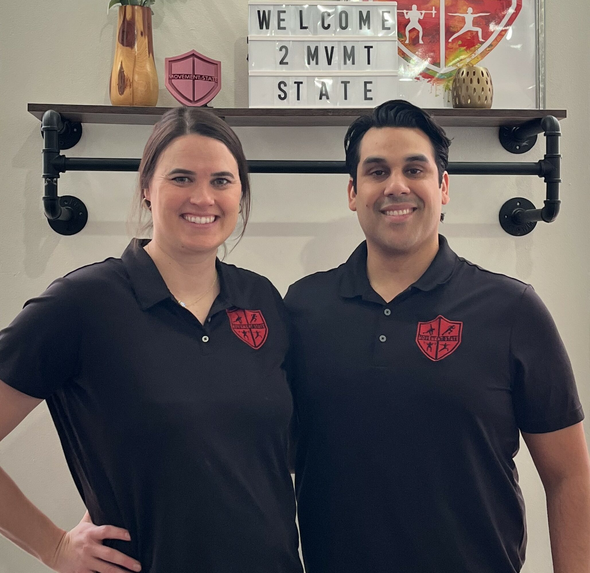 Two people wearing black shirts with red logos stand in front of a shelf with decorative items and a sign that reads 'WELCOME 2 MVMT STATE'.