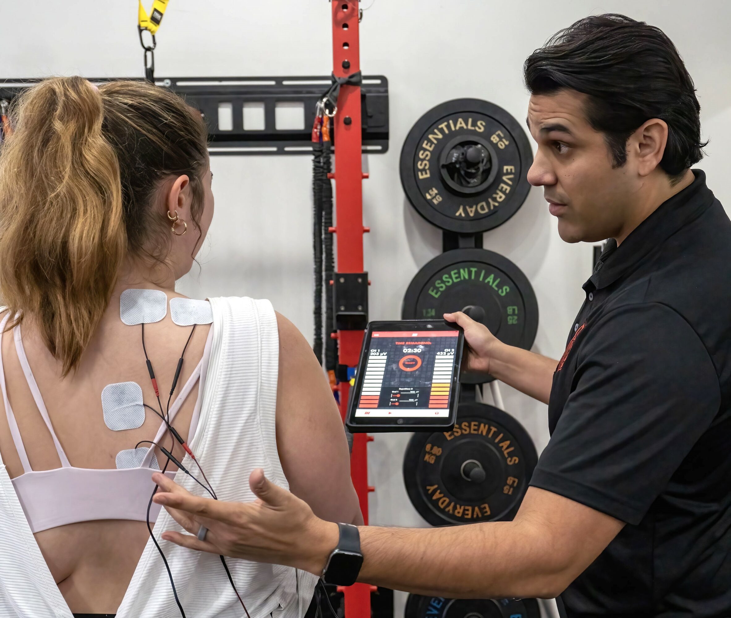 Trainer explains workout data to a woman with electrodes on her back in a gym setting.