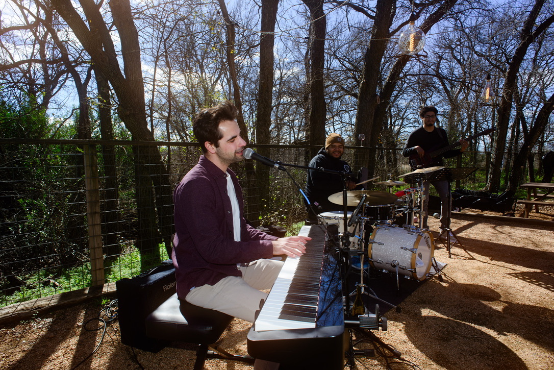 Three musicians playing instruments outdoors in a wooded area, sunlight casting shadows on the ground.