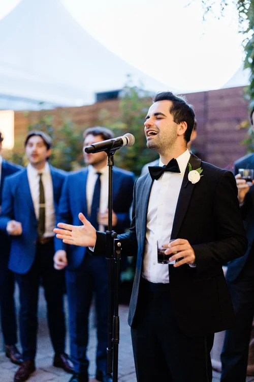 Man in tuxedo speaking into microphone at outdoor event with group of men in suits behind him.