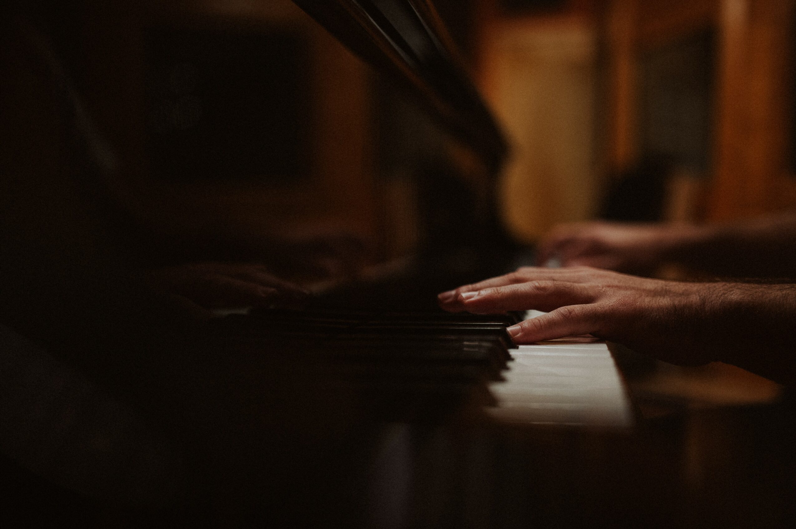 Hands playing piano keys in a dimly lit room, with wooden background and warm lighting.