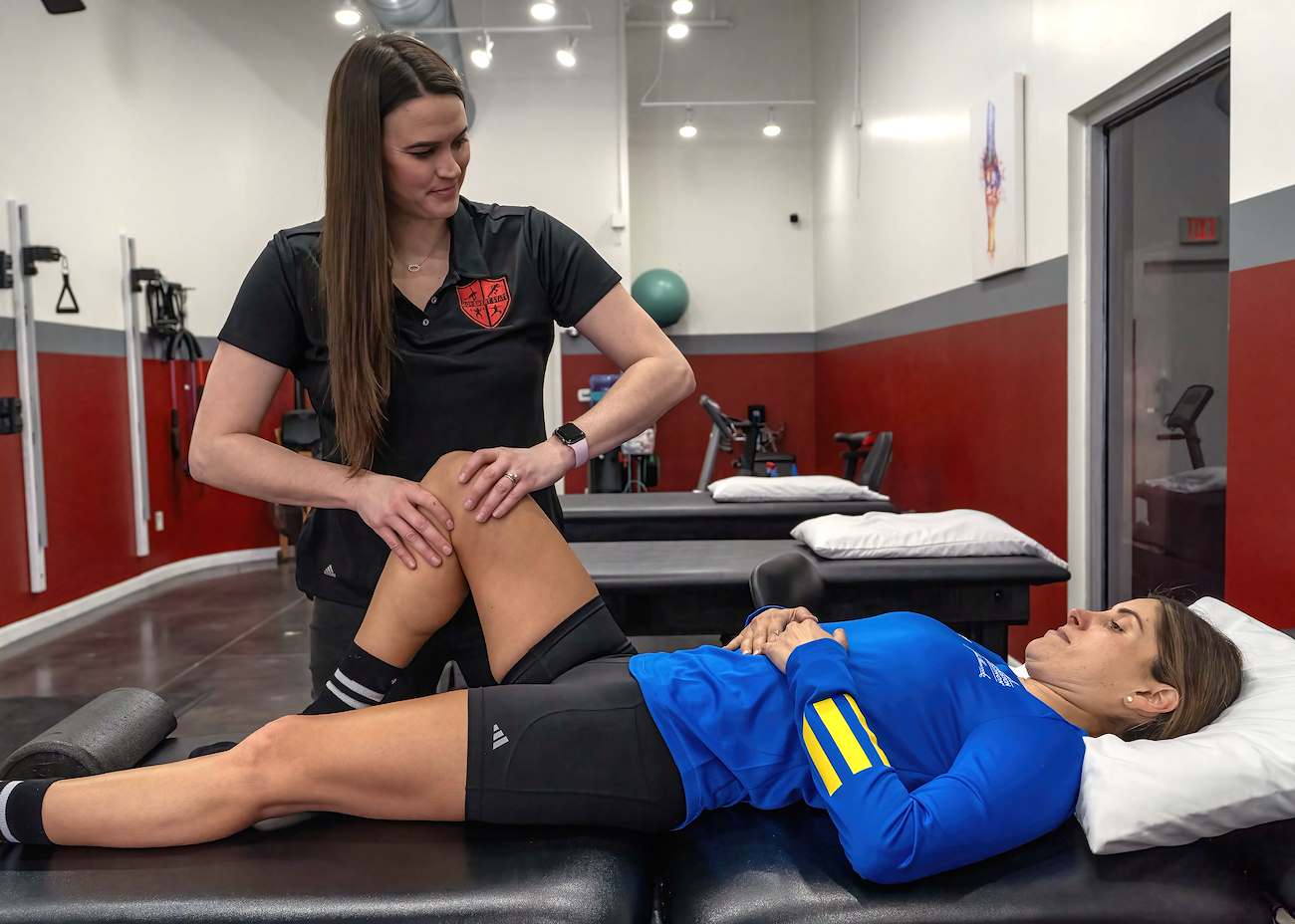 Physical therapist examines a woman's knee while lying on a treatment table in a therapy room.
