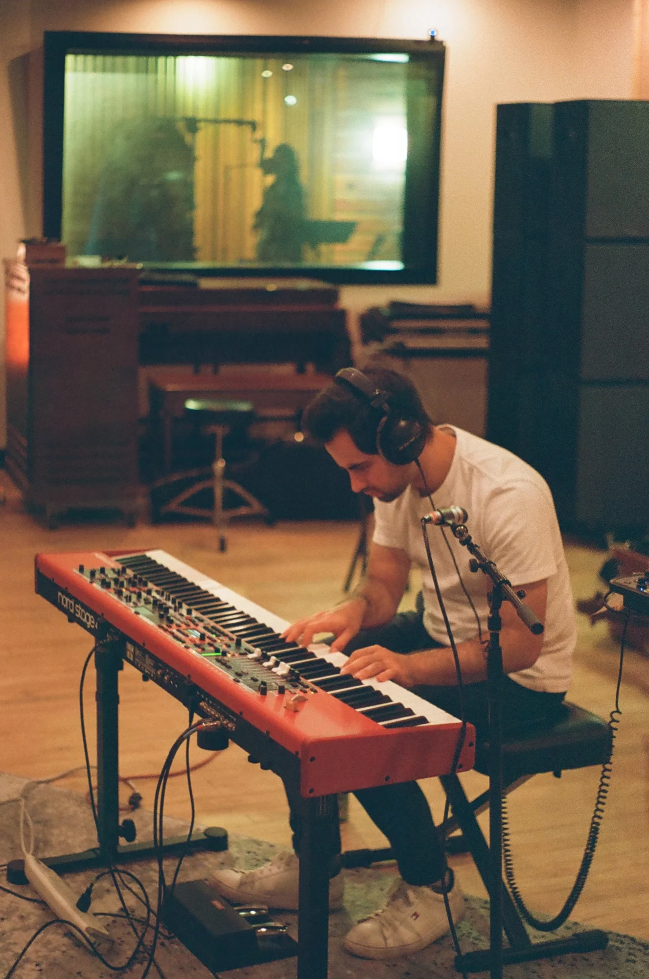 Person wearing headphones playing a keyboard in a recording studio with a large screen and sound equipment.