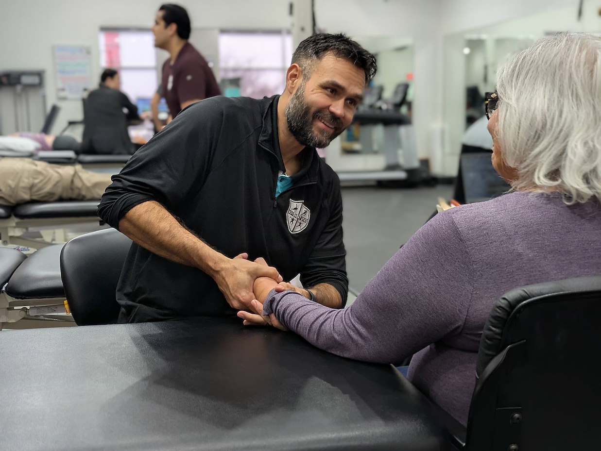Man with dark hair and beard shaking hands with an older woman with gray hair in a medical setting.