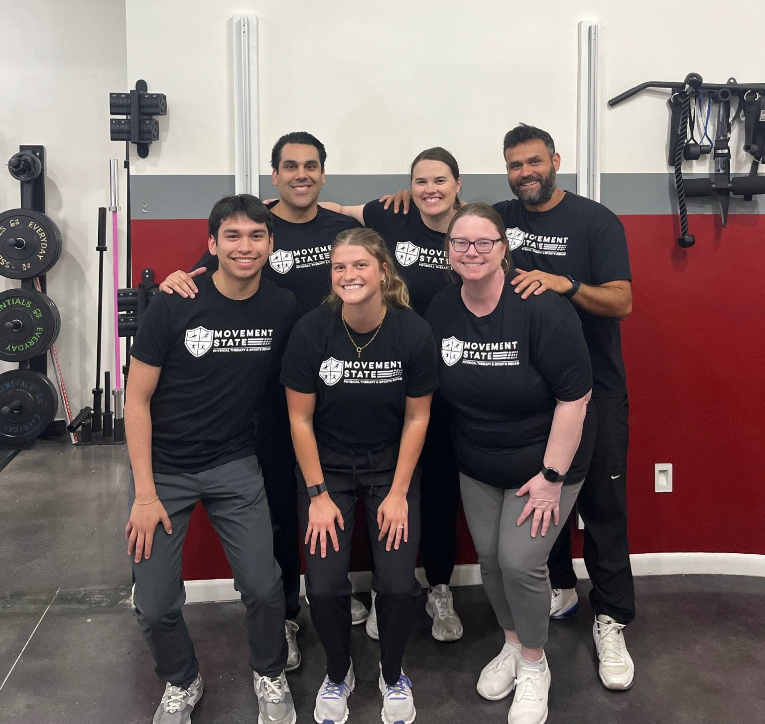Group of six people in a gym, smiling, wearing matching black shirts, standing in front of gym equipment.