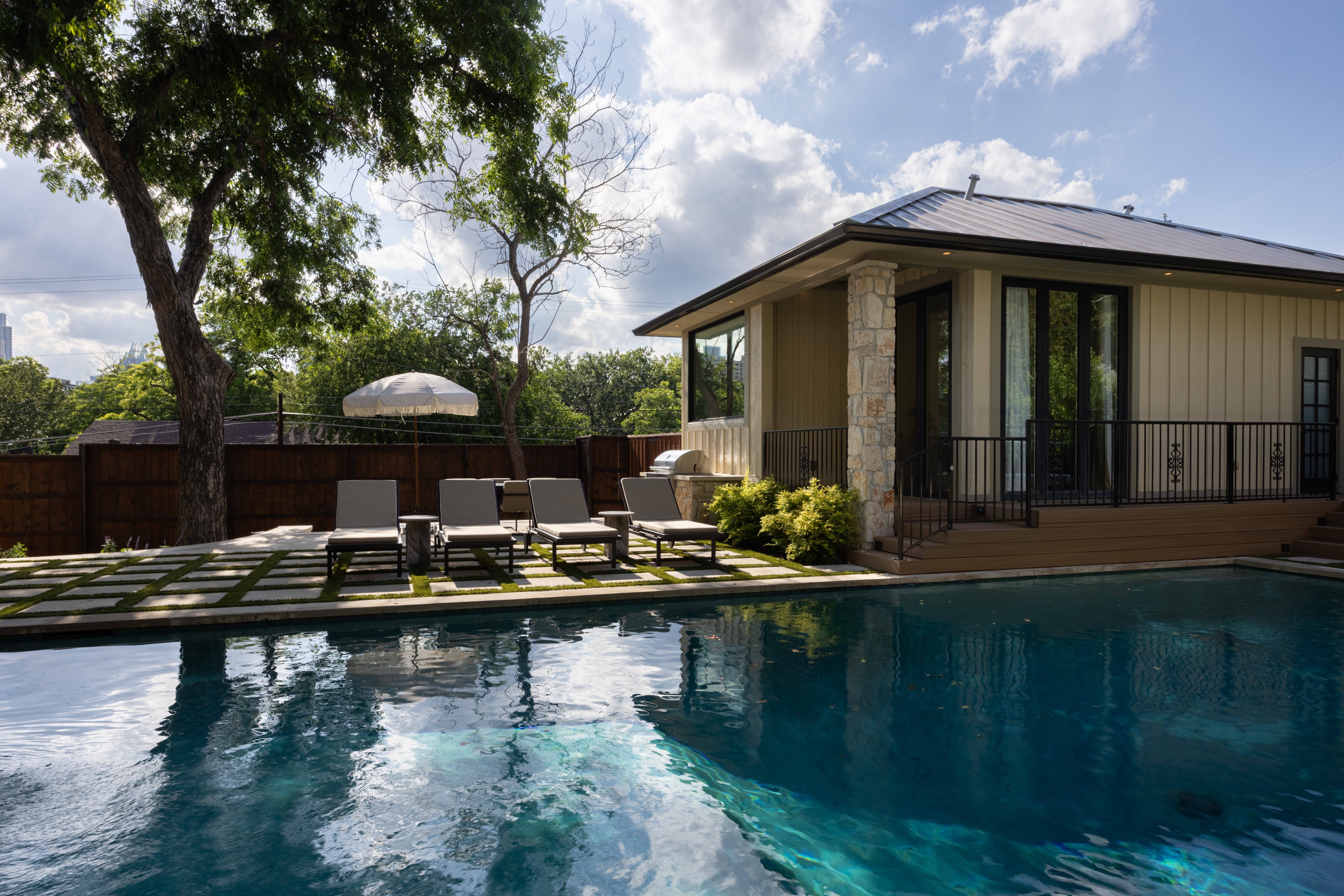 Swimming pool with lounge chairs, a building with large windows, trees, and a blue sky with clouds.
