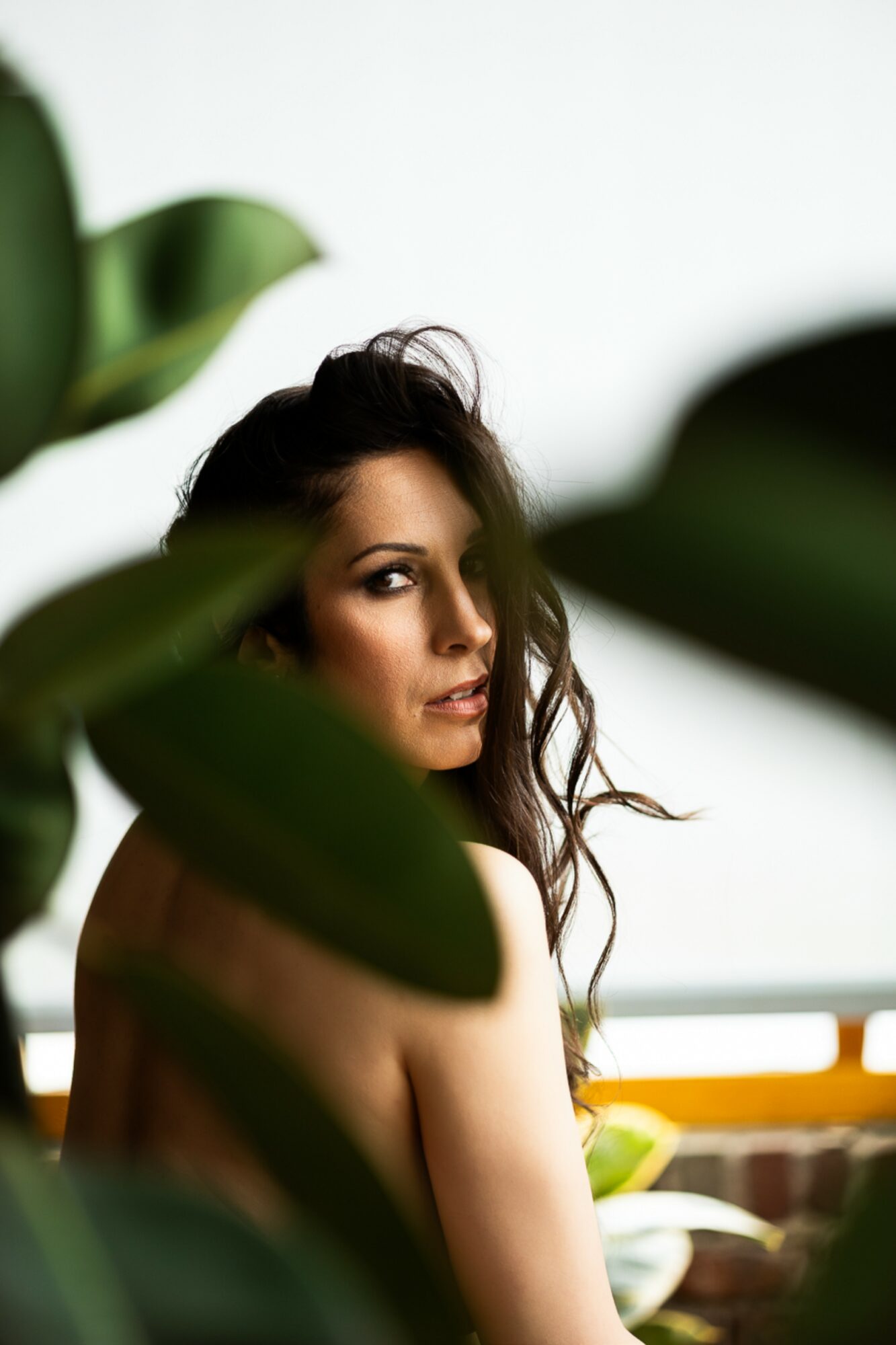 A woman with long, wavy hair looks over her shoulder through green leaves, with a blurred background and natural lighting.