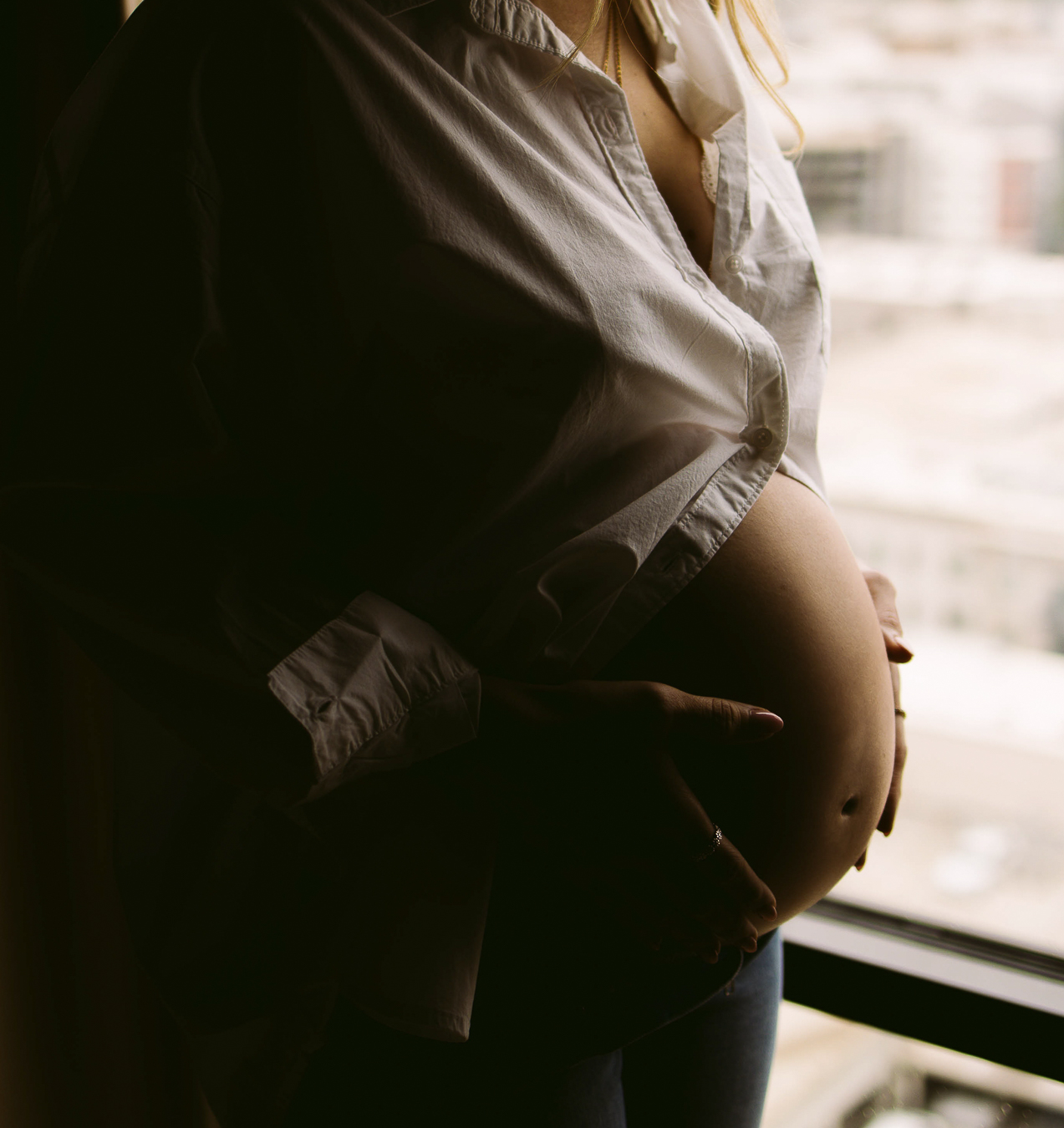 Pregnant woman standing near window, hand on belly, cityscape background, soft lighting, casual clothing.