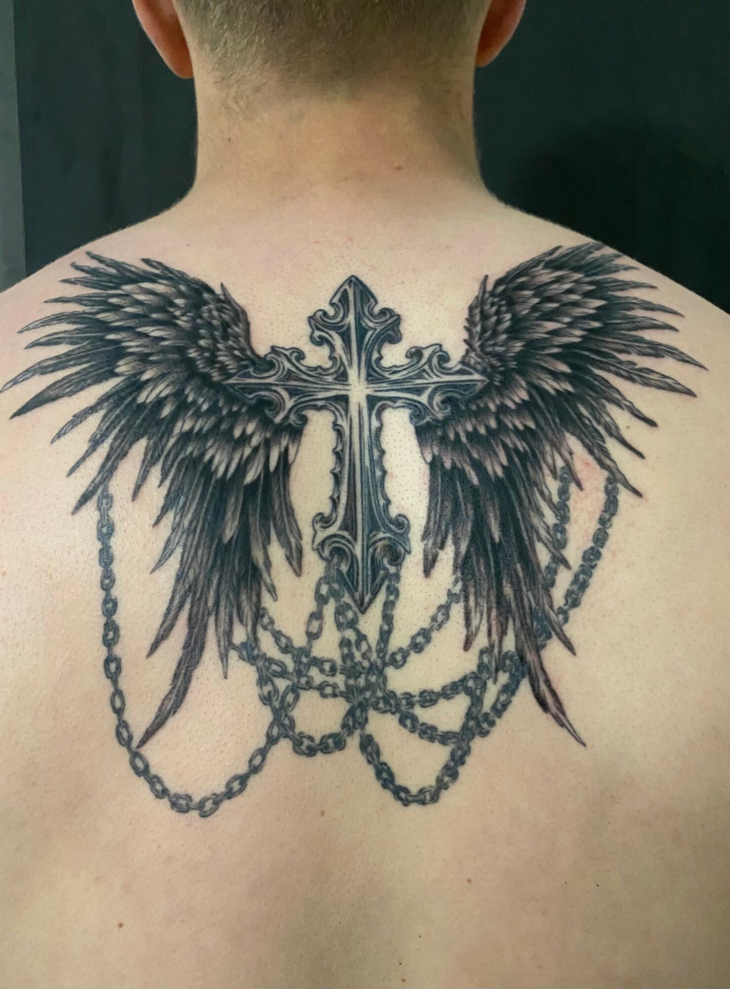Back tattoo of a cross with angel wings and hanging chains, detailed and symmetrical, covering upper back.