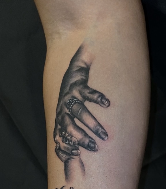 Black and gray tattoo of a hand with rings, fingers pointing downward on person's arm.