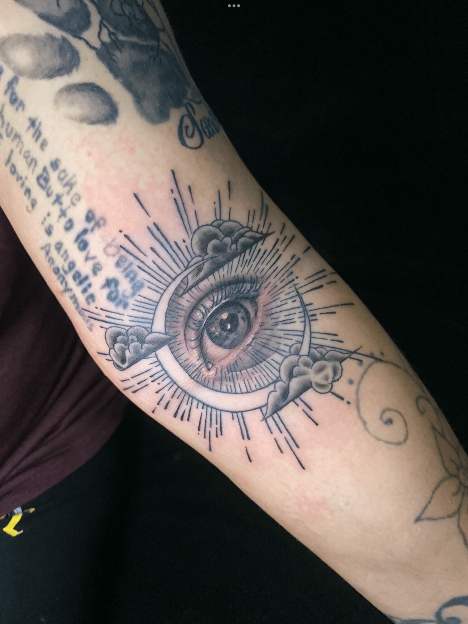 Tattoo of an eye with rays, clouds, and decorative elements on a person's arm.