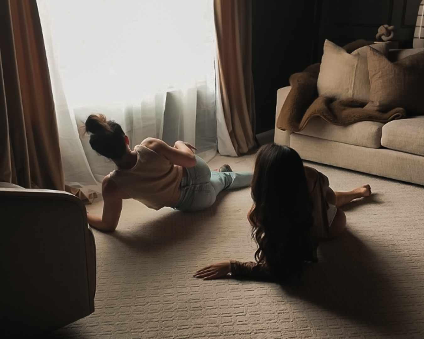 Two people sit on the floor near a window with curtains, facing away from the camera, in a living room.