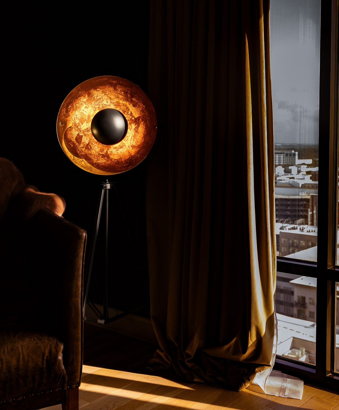 A moon-shaped lamp with a black sphere in the center stands near a window with curtains, cityscape outside, warm indoor lighting.