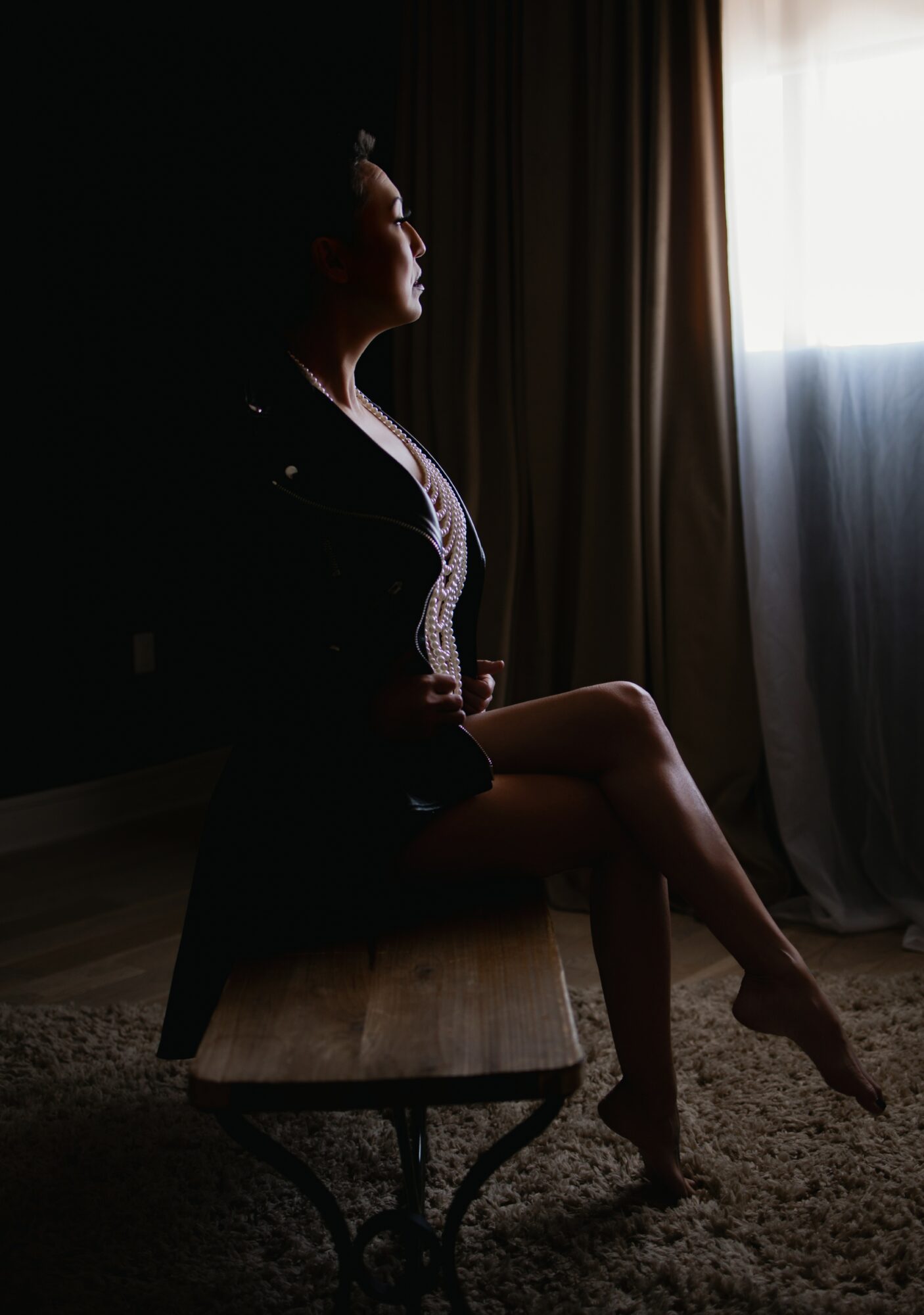 Woman sitting on a bench near a window with curtains, looking outside, with light illuminating her profile.