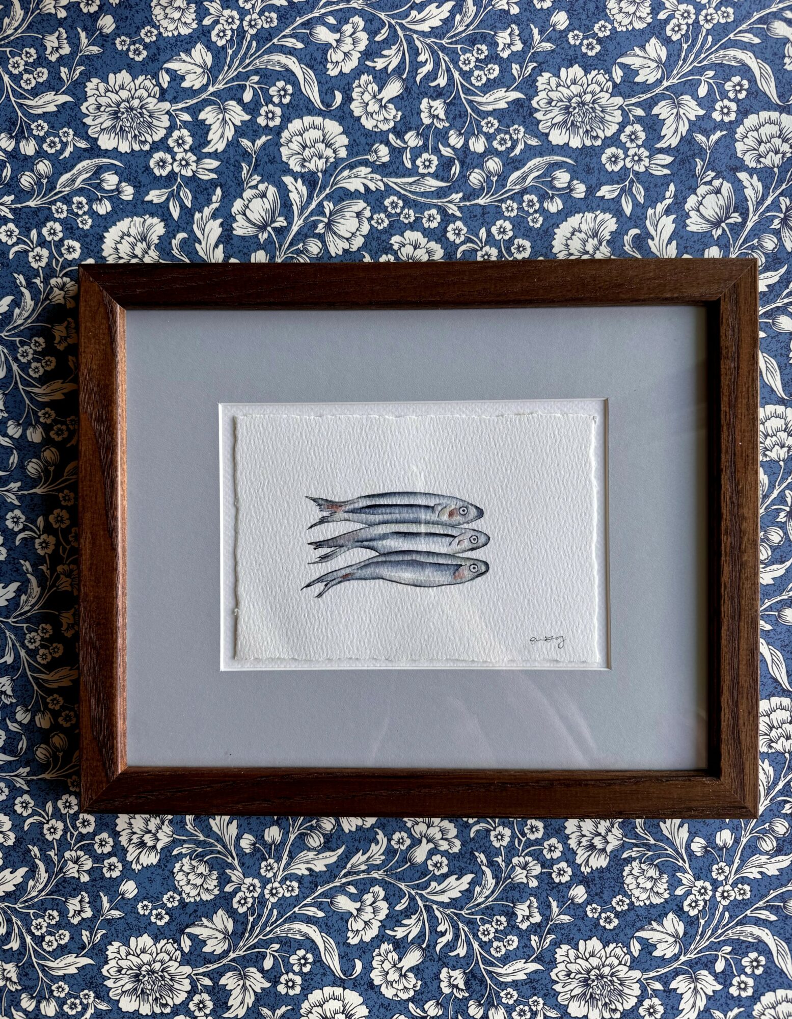 Framed artwork of three fish swimming, on a blue floral patterned background.