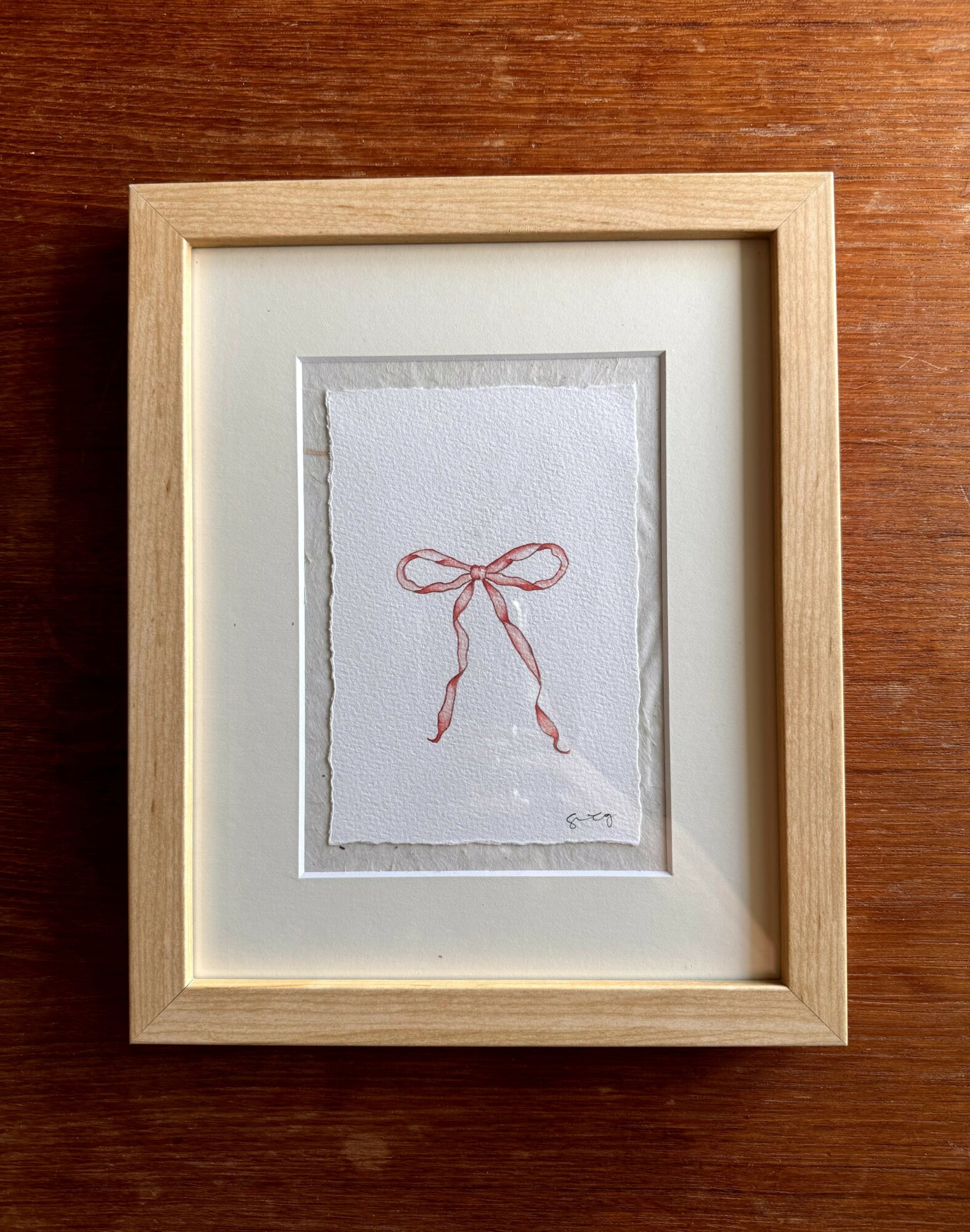 Framed artwork with a simple red bow illustration on white paper, placed on a wooden surface.