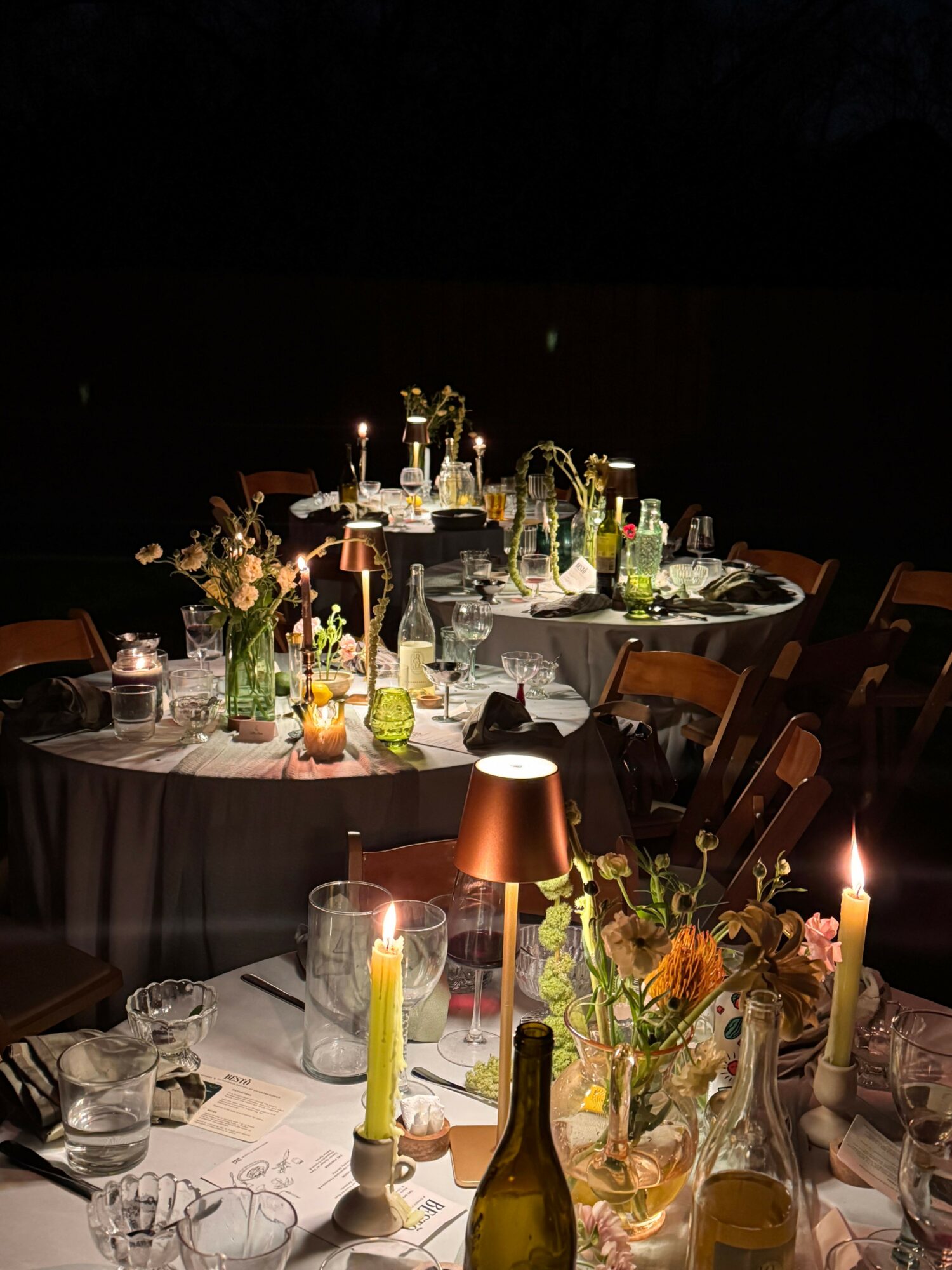 A table set with bottles, candles, flowers, and glasses for a formal event, viewed from the side.