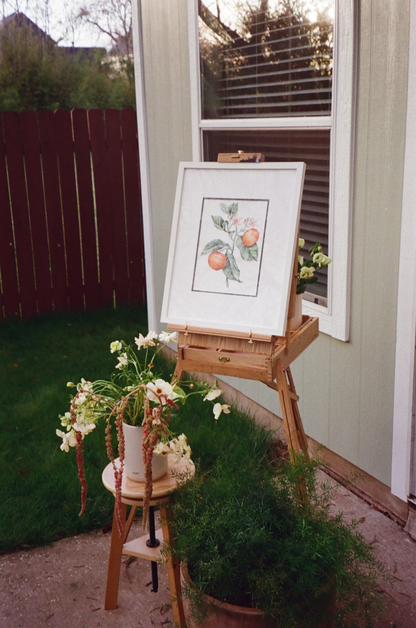 Framed artwork on a wooden easel outside a house, with a potted plant and garden in the background.