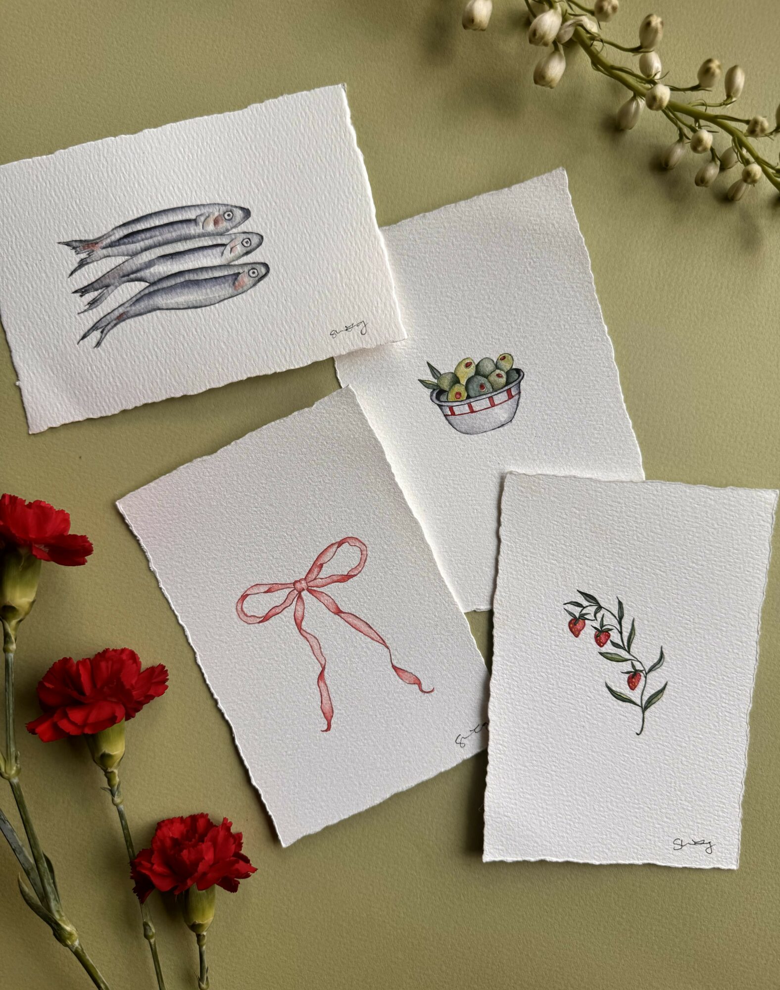 Four small watercolor paintings on paper, surrounded by red flowers and a sprig of white berries, on a green surface.