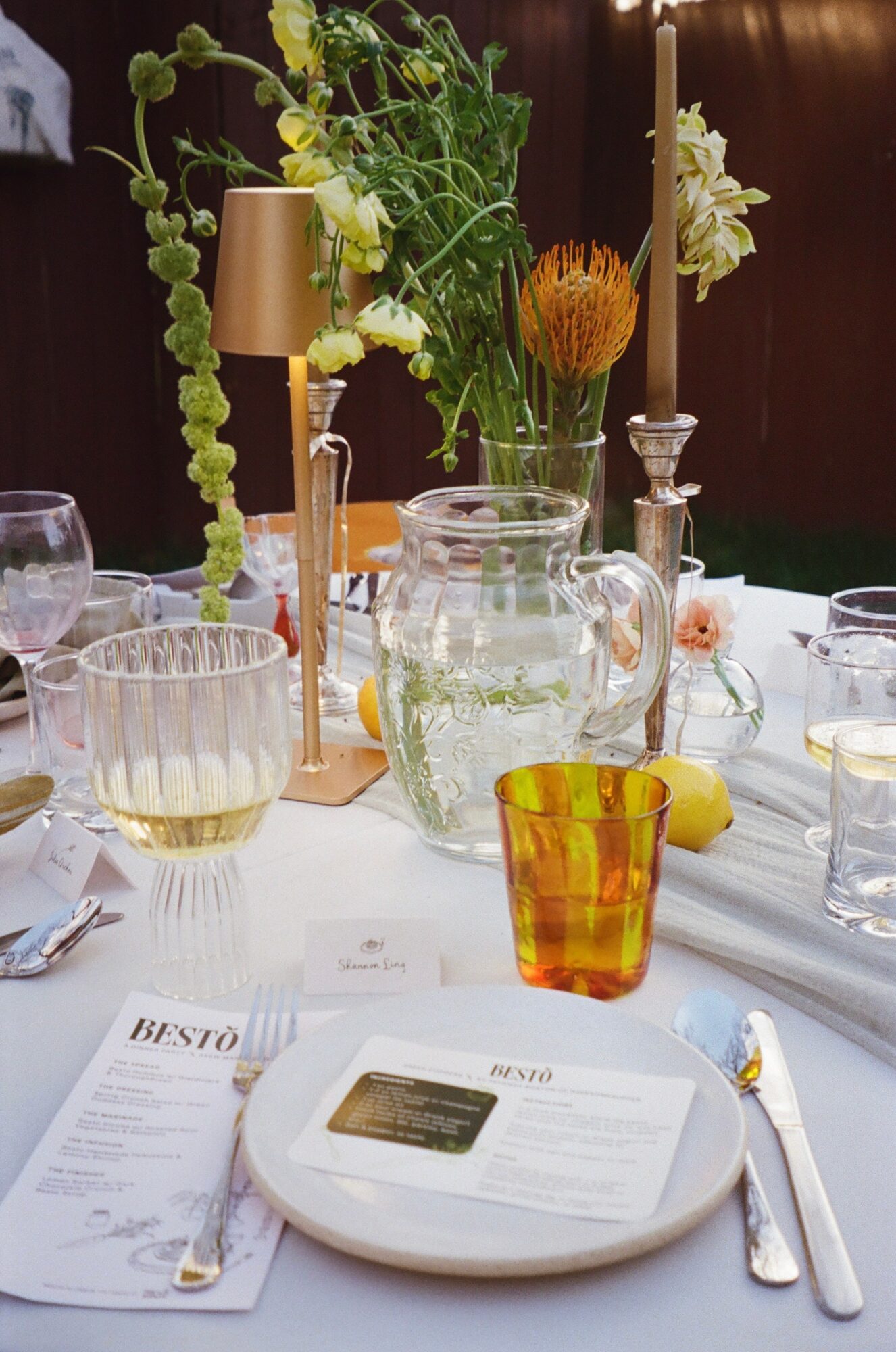 Table setting with flowers, glasses, a pitcher, and a yellow-orange glass, with a plate and utensils in foreground.