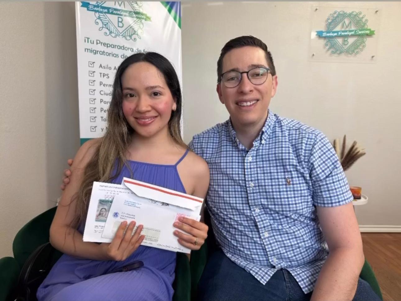 Two people sitting indoors, smiling, holding a document or certificate, with a banner and wall decorations in the background.