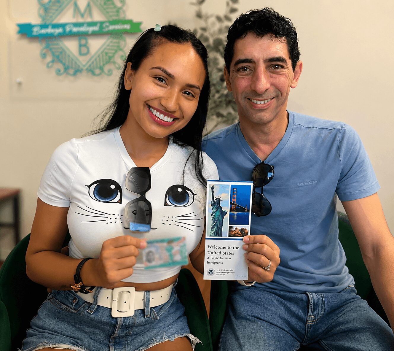 Two people smiling, holding a brochure and a small plush toy, sitting indoors with a sign in the background.