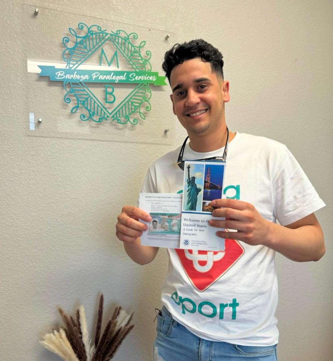 Young man smiling, holding pamphlet, standing in front of a sign on a wall, wearing a white t-shirt with a red and blue logo.