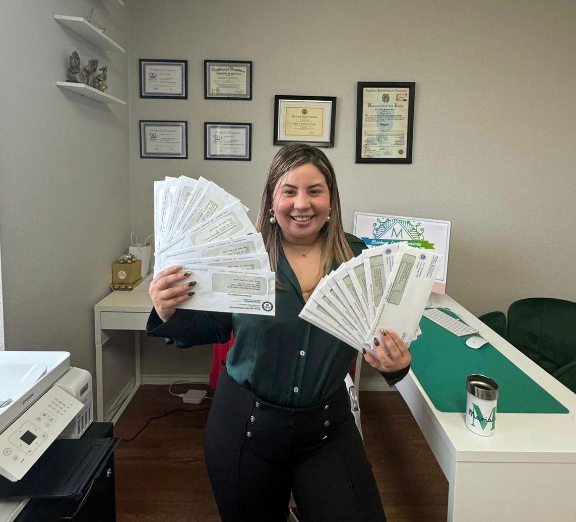 Woman smiling holding multiple certificates in an office, certificates on wall behind her, desk with papers and a cup nearby.