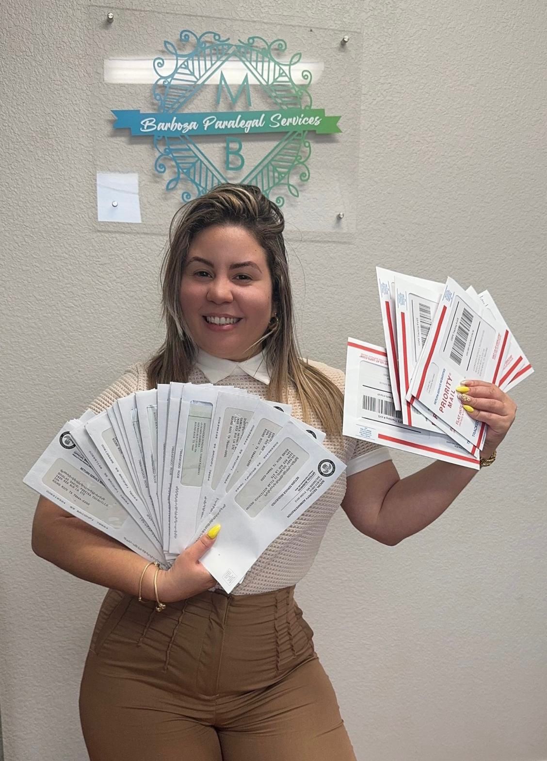 Young woman smiling, holding multiple envelopes or documents in both hands, standing indoors in front of a wall with a sign.