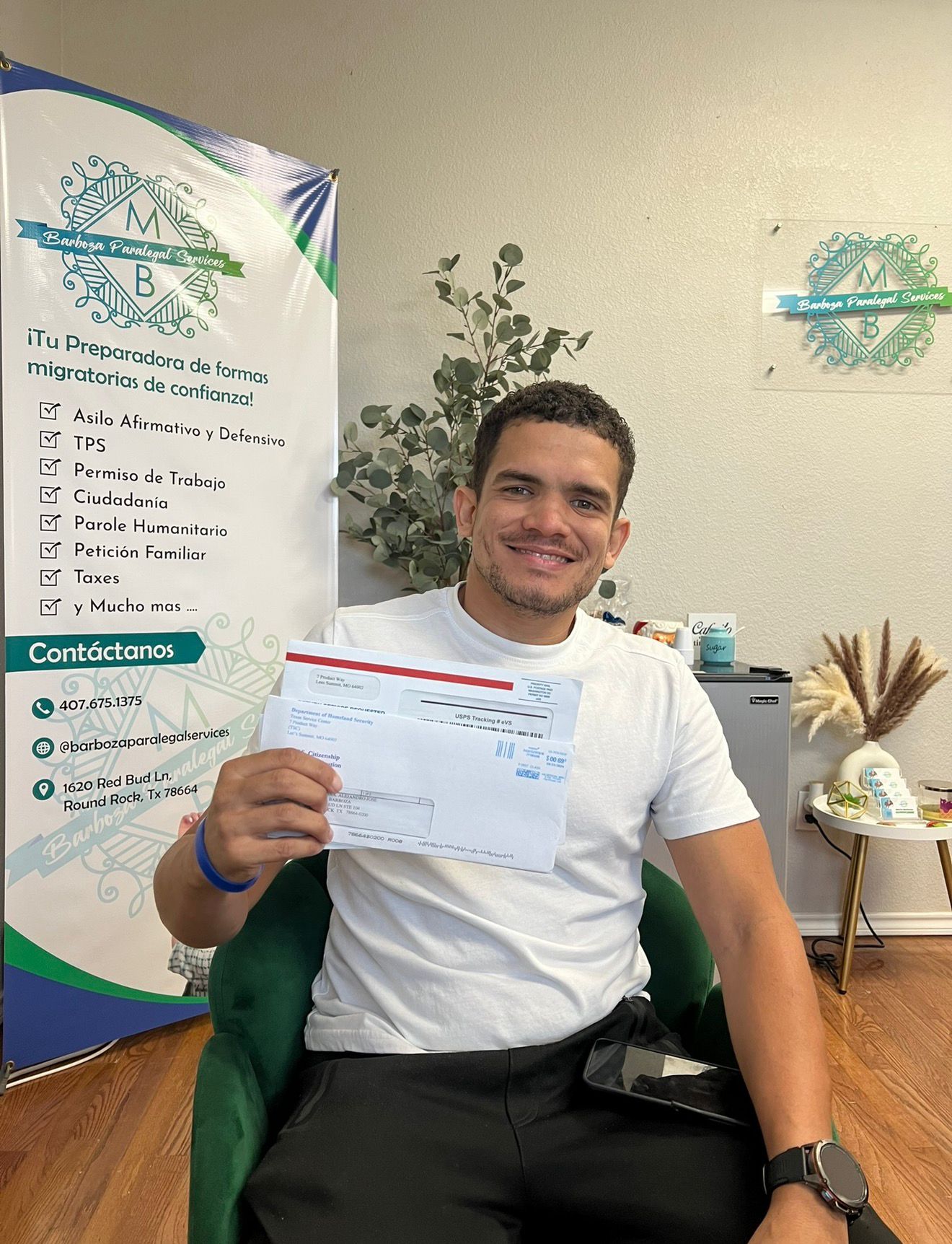 Man sitting in a chair holding a document, smiling, with a banner and plant in the background.