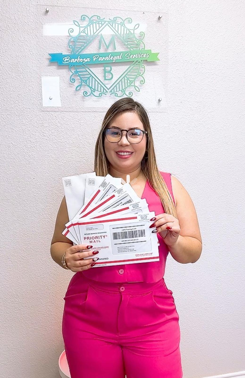 Woman with glasses holding multiple flyers, smiling, standing in front of a wall with a logo, wearing a pink dress.