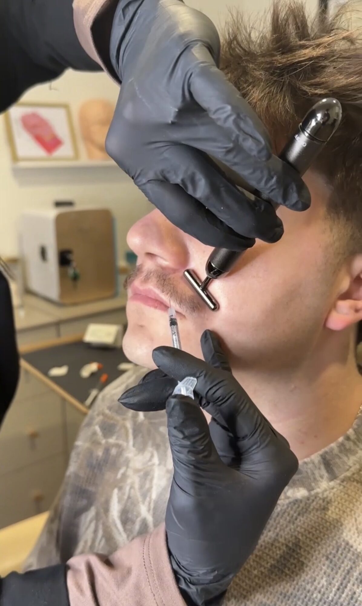 Person receiving a nose piercing, wearing black gloves, in a clinical setting with equipment in background.
