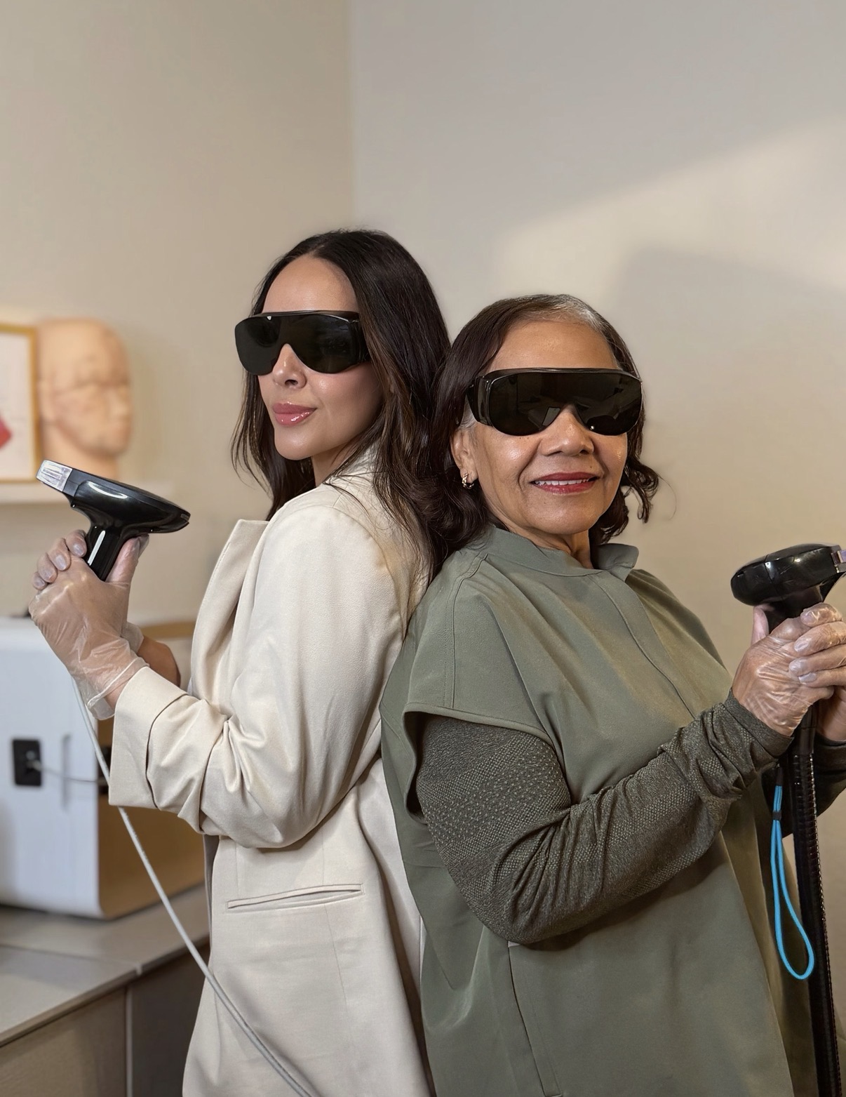 Two women wearing sunglasses and holding gaming controllers, standing back-to-back indoors, smiling. One woman has long dark hair, the other has short dark hair.